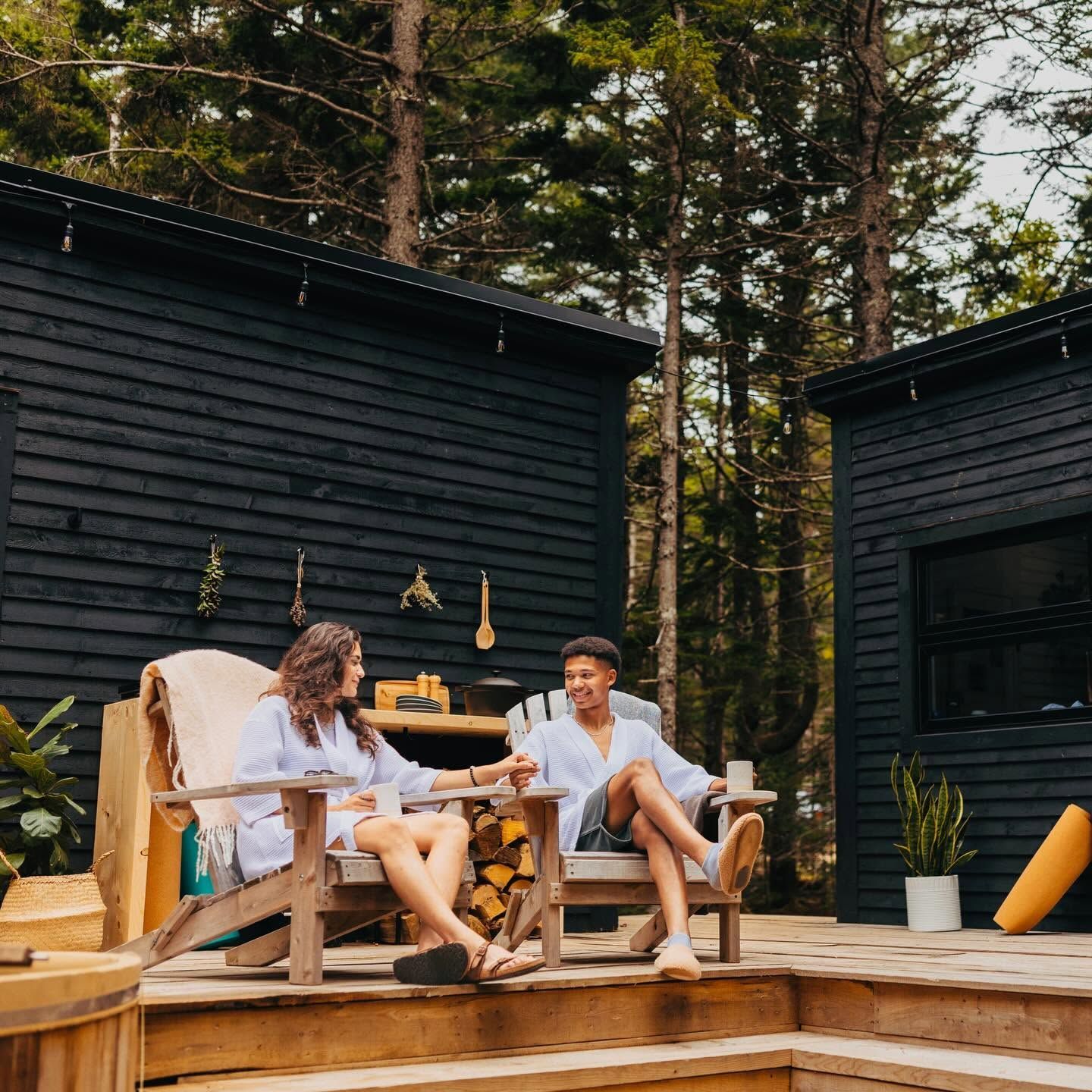 Twee mensen zitten op houten stoelen bij een zwarte hut en praten op een zonnig terras.