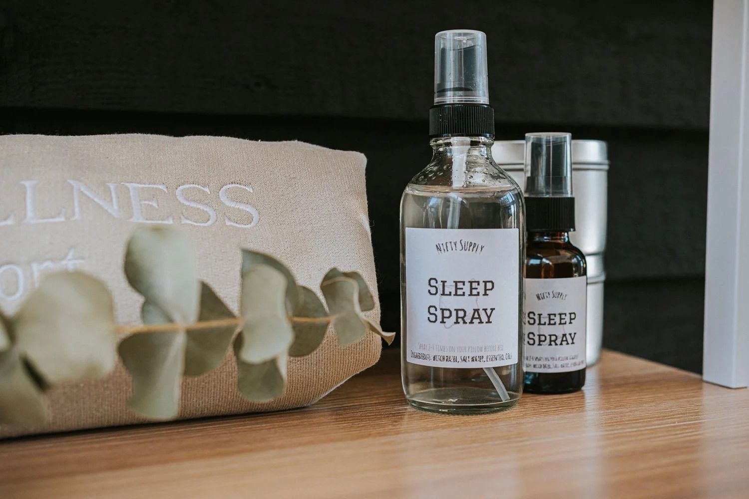 Slaapsprayflesjes op een houten tafeltje naast een beige wellness-tasje en groene bladeren.