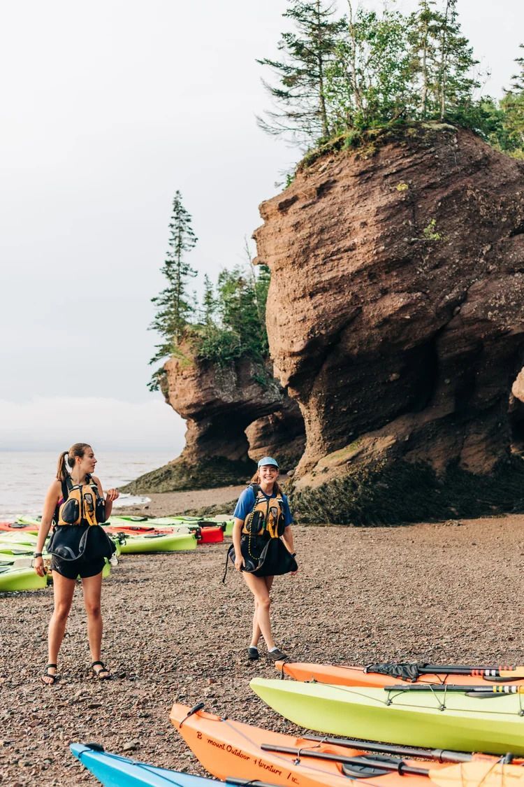 Twee wandelaars bij kleurrijke kajaks op een rotsachtig strand onder een grote klif met bomen.