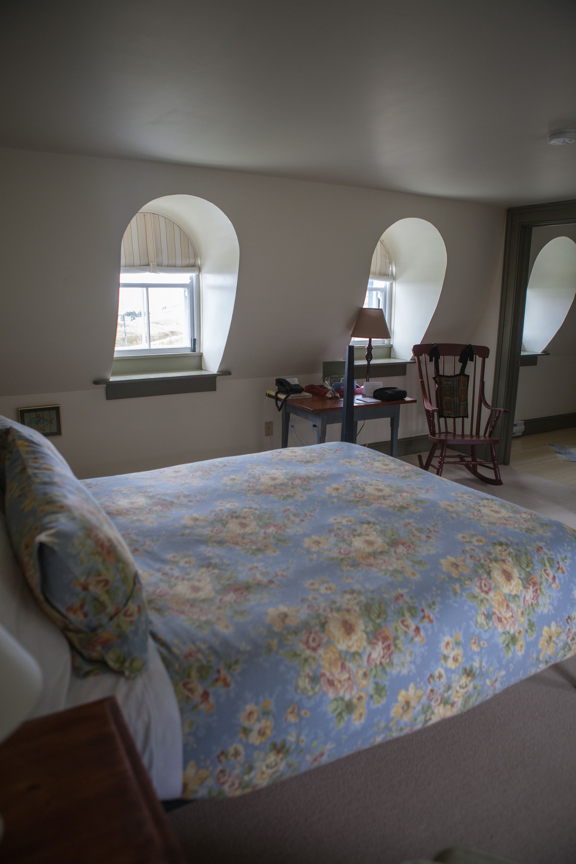 Slaapkamer van Fisher's Loft Inn in Port Rexton met een blauw bloemenpatroon op het bedsprei, boogvensters en een houten schommelstoel tegen de muur.