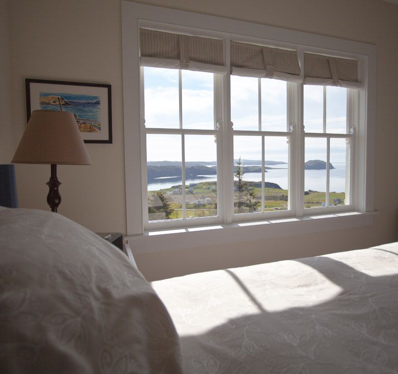 Zonnige slaapkamer van Fisher's Loft Inn in Port Rexton met wit bed, groot raam en uitzicht op de oceaan en het eilandlandschap.
