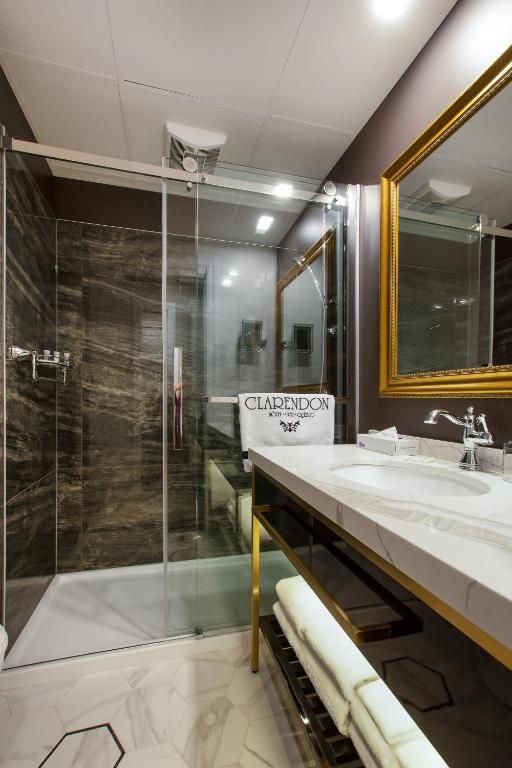 Moderne badkamer met glazen douchecabine, marmeren tegels, witte wastafel en spiegel met gouden frame.