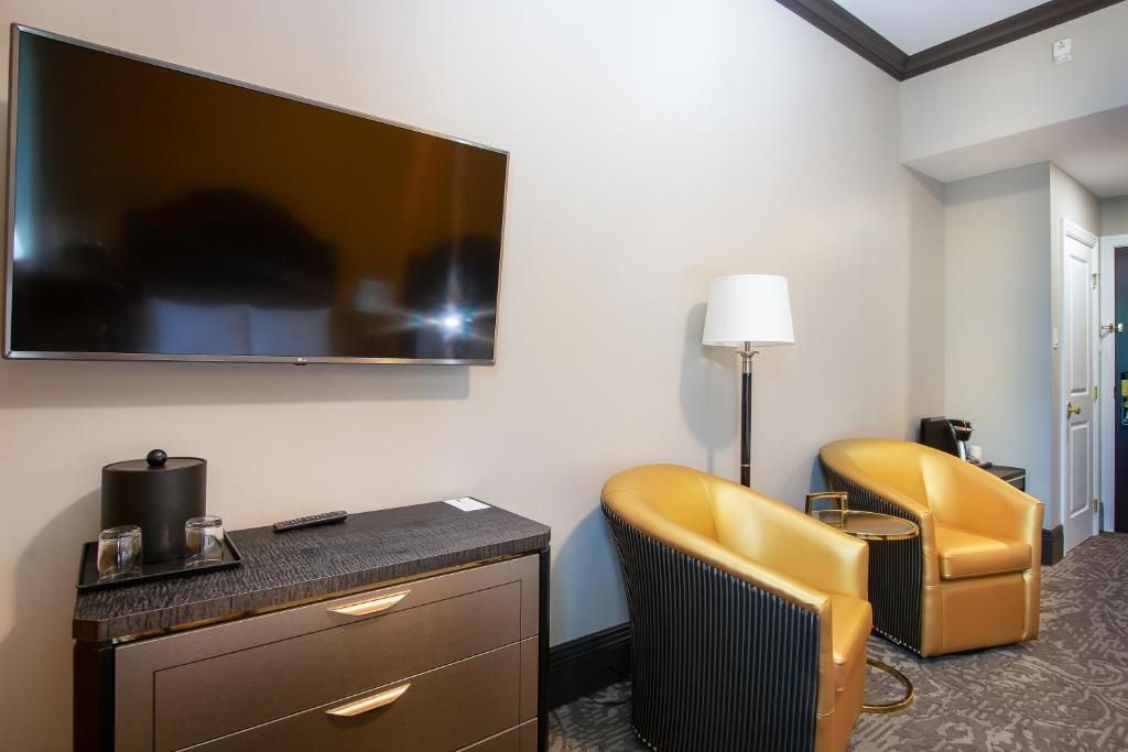 Hotelkamer met aan de muur gemonteerde tv, koffiezetapparaat en twee gele fauteuils bij een staande lamp.