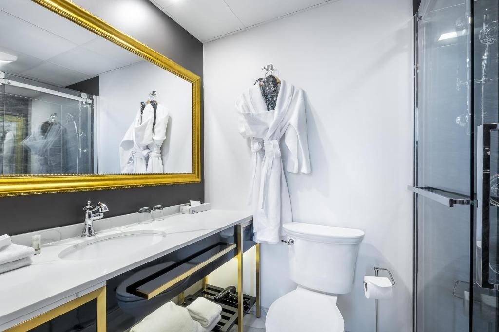 Moderne witte badkamer met goudkleurige spiegel, badjas aan de muur, wastafel, toilet en glazen douchedeur.