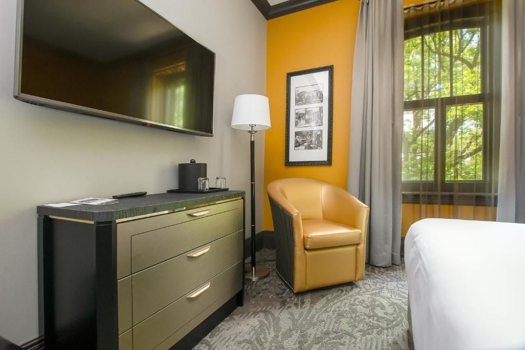 Hotelkamer met gele accentmuur, fauteuil, commode, aan de muur gemonteerde tv en raam met gordijnen.