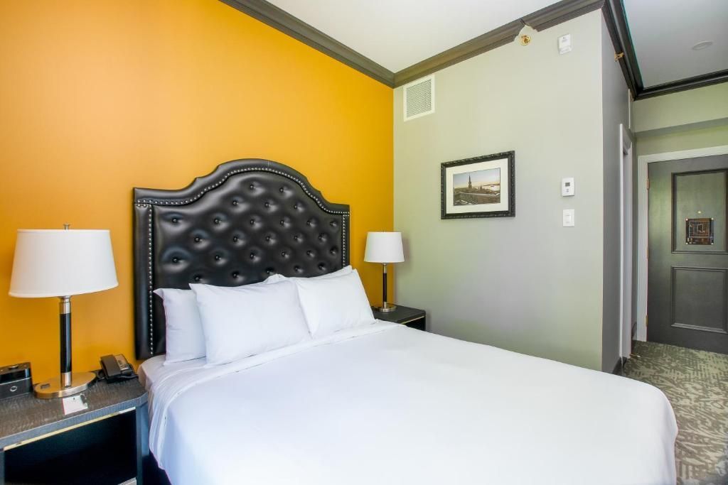 Hotelkamer met een netjes opgemaakt bed, een donker gecapitonneerd hoofdeinde, een gele accentmuur en nachtlampjes.