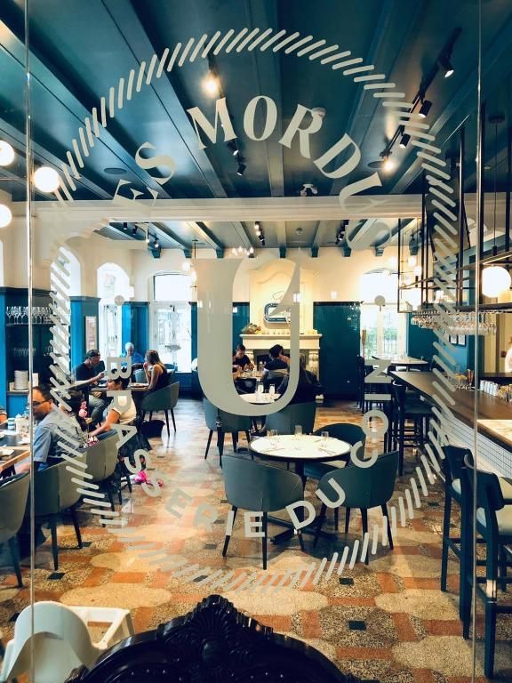 Druk caféinterieur met zittende klanten, een glazen boogvormige ingang en een vloer met decoratieve tegels.