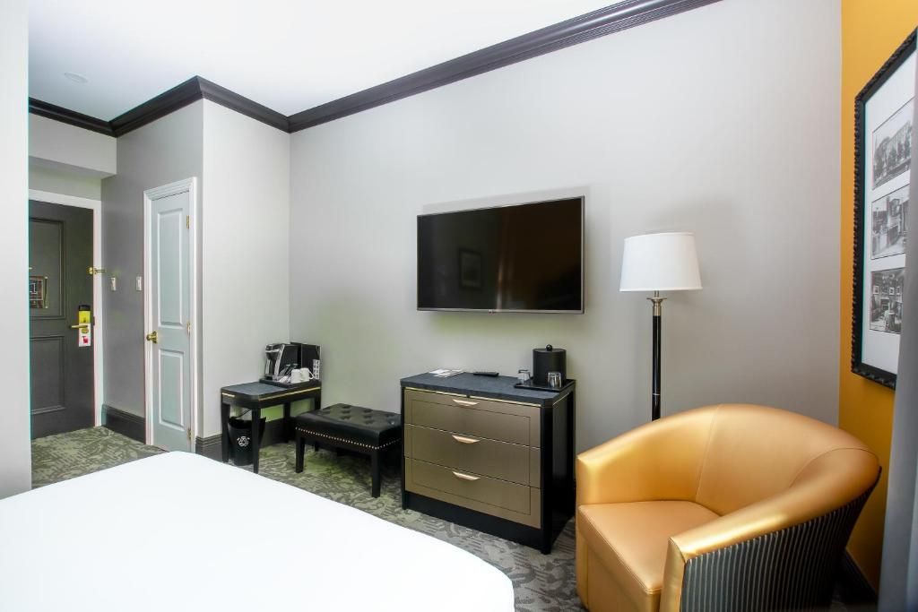 Hotelkamer met tv, lamp, bureau en beige stoel naast het bed.