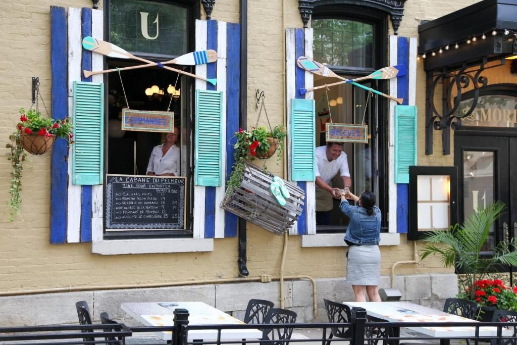 Twee open ramen met blauwe luiken, met daarbuiten een mand die buiten op een terras van een café hangt.