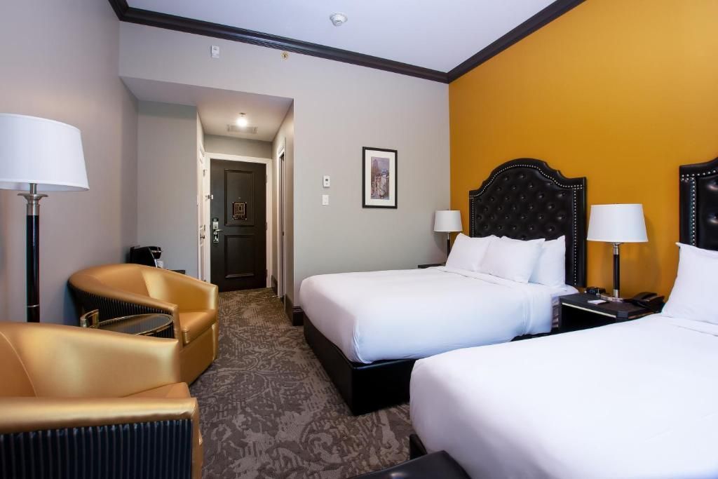 Hotelkamer met twee bedden, een gele accentmuur, beige stoelen en een deur naar de gang.