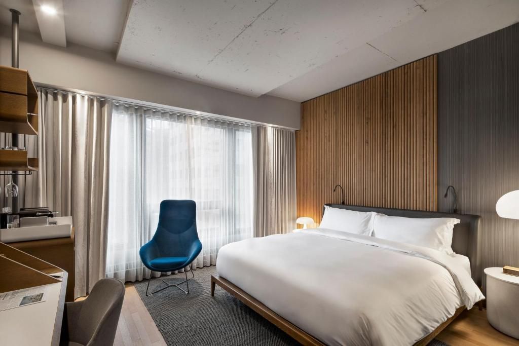 Moderne hotelkamer van Hôtel Le Germain Montréal met een groot bed, een blauwe stoel, een houten lambrisering en transparante gordijnen voor het raam.