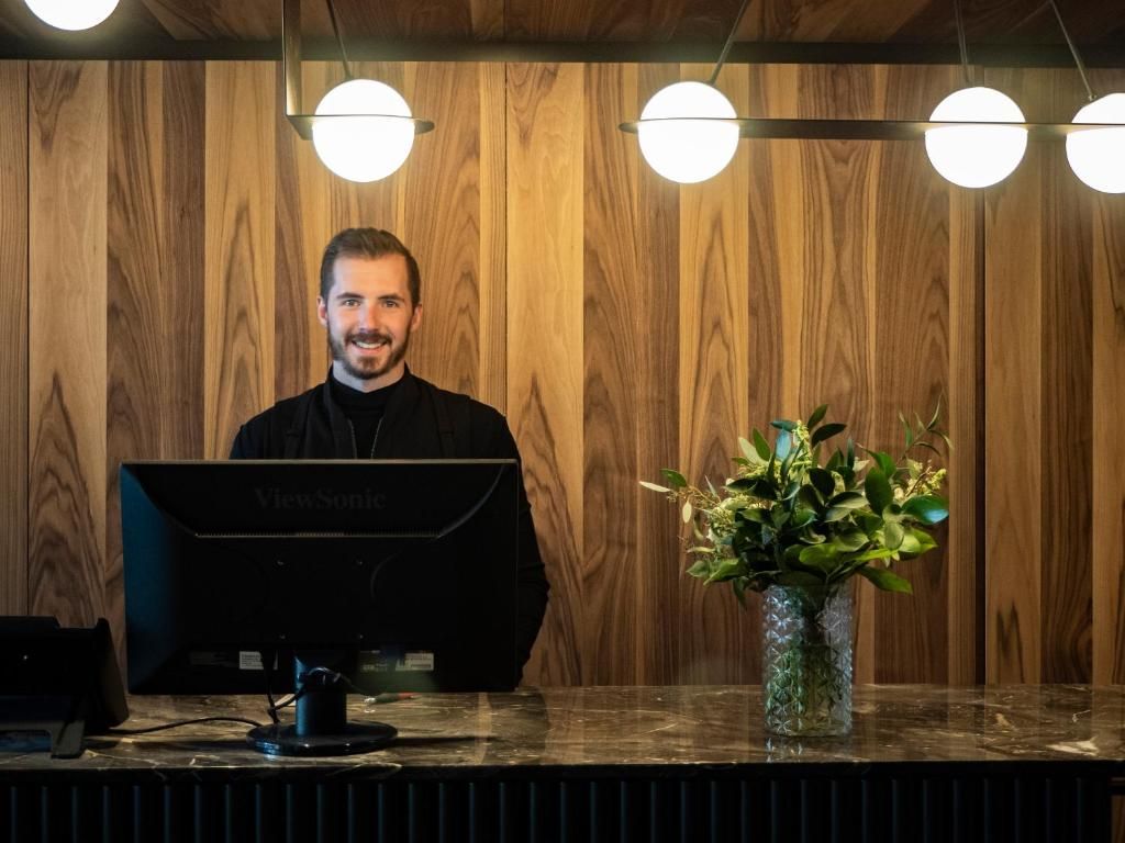 Een glimlachende receptioniste van Hôtel Le Germain Montréal zit achter een bureau met een monitor en bloemen in een lobby met houten lambrisering.
