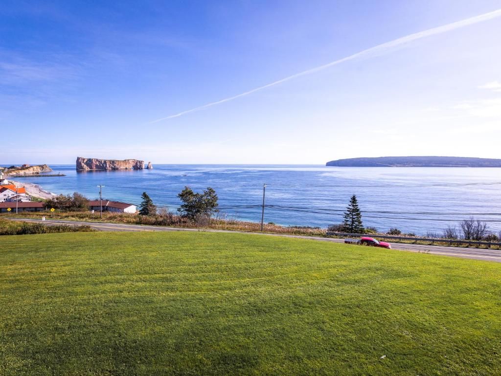 Een grasrijk park aan zee in Percé, Canada, met uitzicht op kalm blauw water, een rotsachtige kustlijn en eilanden in de verte onder een heldere hemel.