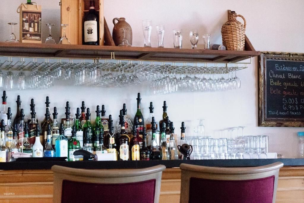 Een barplank met diverse flessen sterke drank, hangende glazen en barkrukken in een gezellige bar van Hotel Tadoussac.