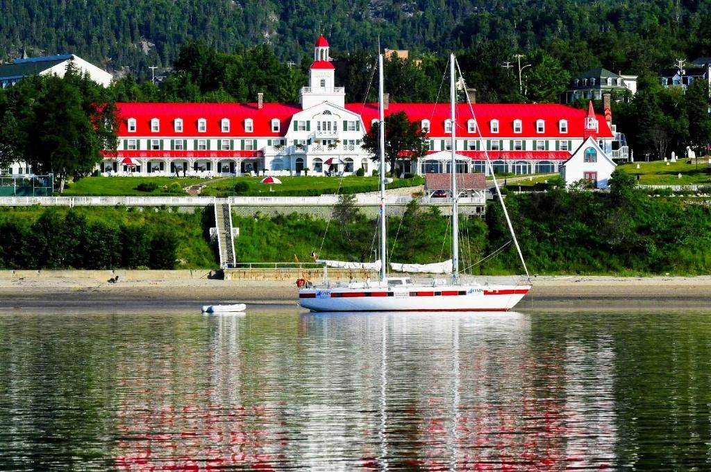 Zeilboot op kalm water voor Hotel Tadoussac met een rood dak aan de waterkant en een beboste heuvel.