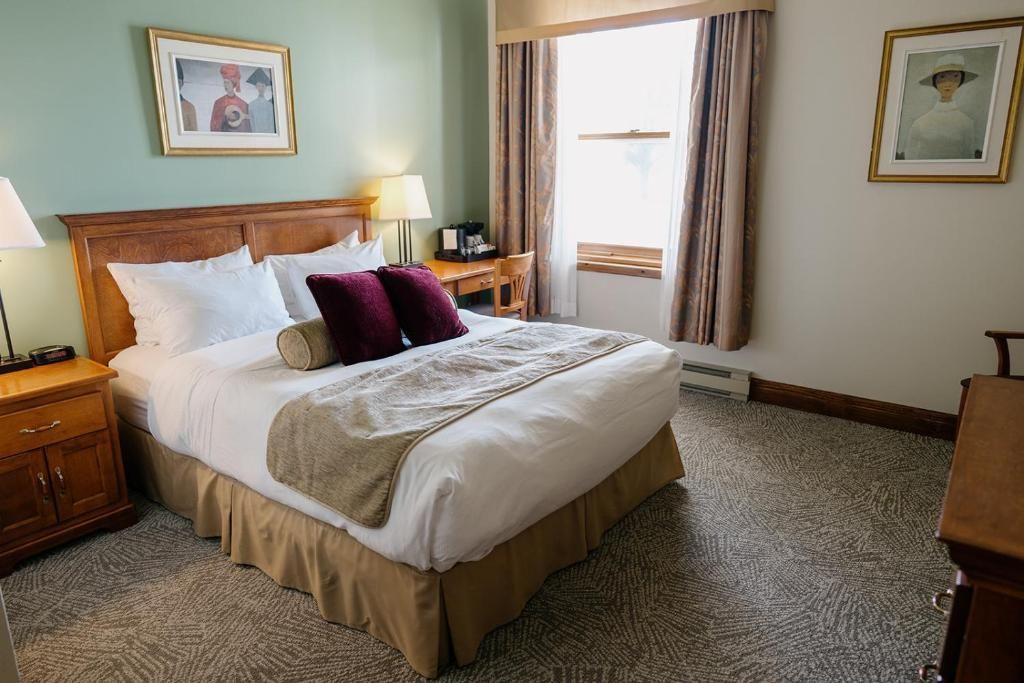 Gezellige hotelkamer van Hotel Tadoussac met een houten bed, wit beddengoed, paarse kussens en een raam met gordijnen.