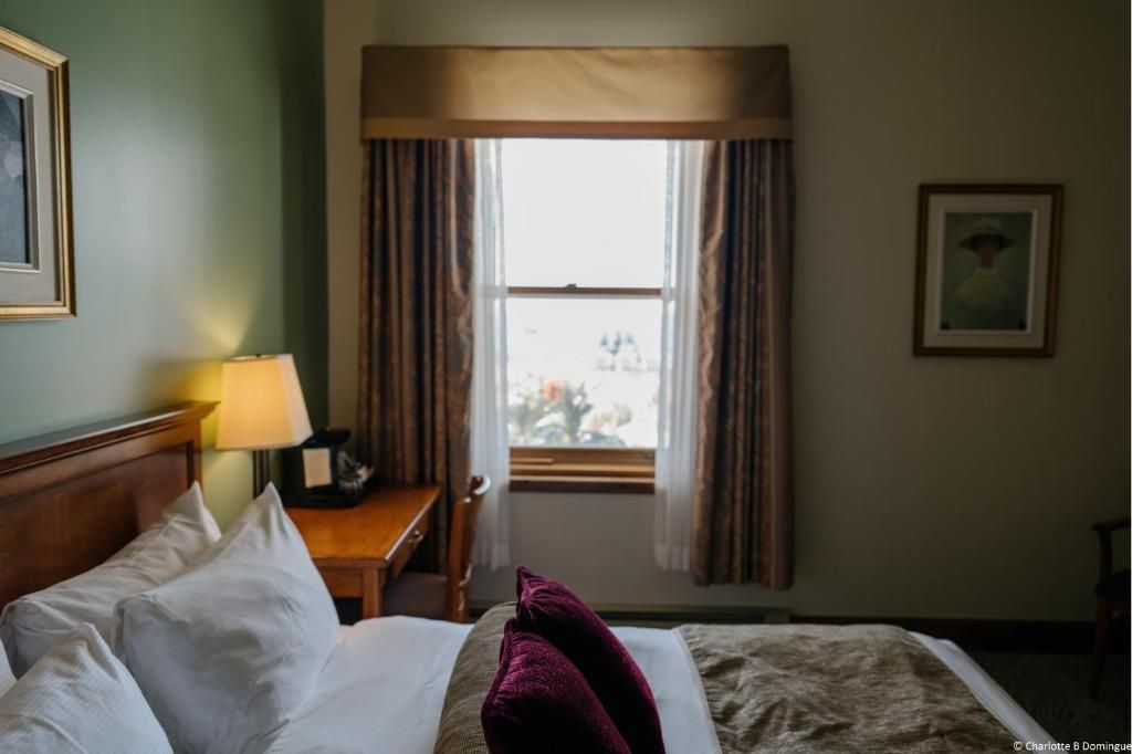 Hotelkamer van Hotel Tadoussac met opgemaakt bed, nachtlampje en raam met gordijnen.