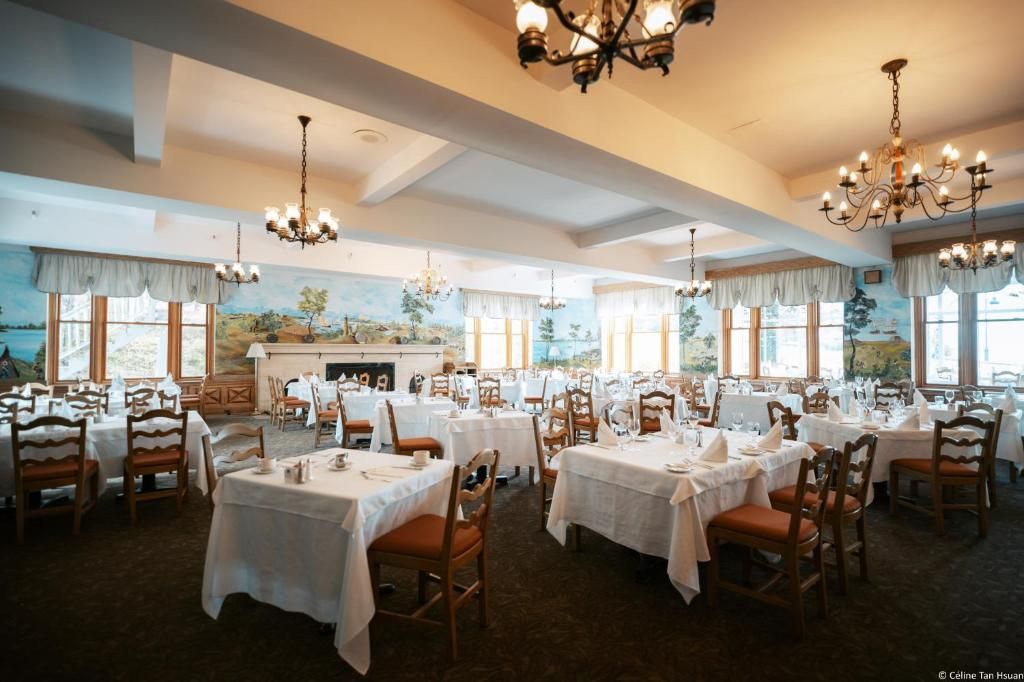 Elegante feestzaal van Hotel Tadoussac met ronde tafels, houten stoelen, kroonluchters en ramen met uitzicht op de oceaan.