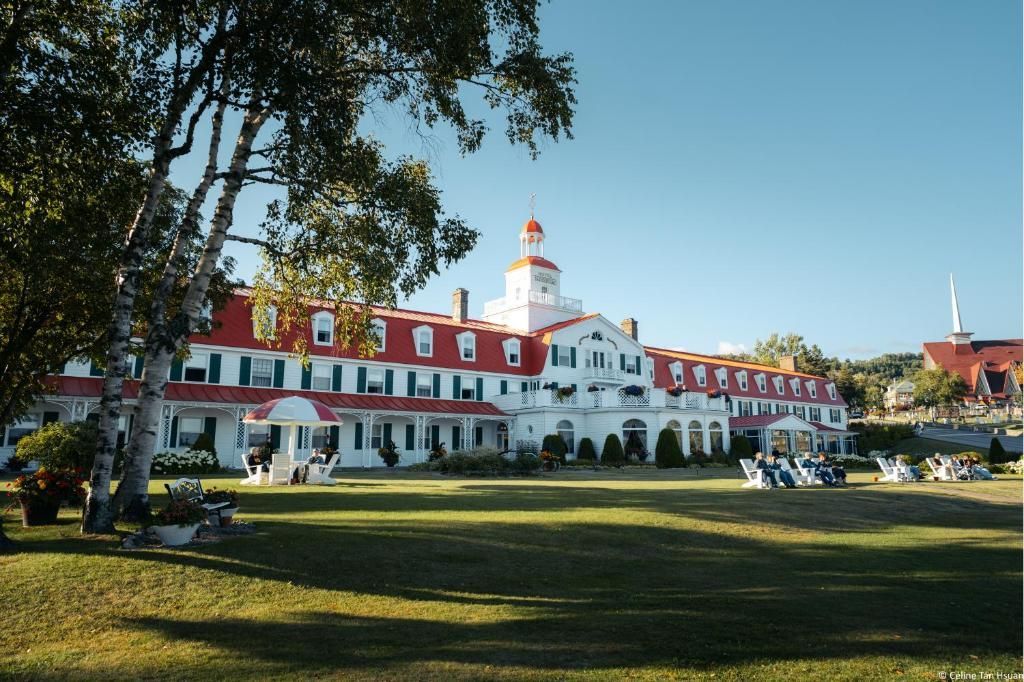 Groot wit-rood koloniaal gebouw van Hotel Tadoussac met een toren, gelegen achter een grasveld en bomen onder een helderblauwe hemel.