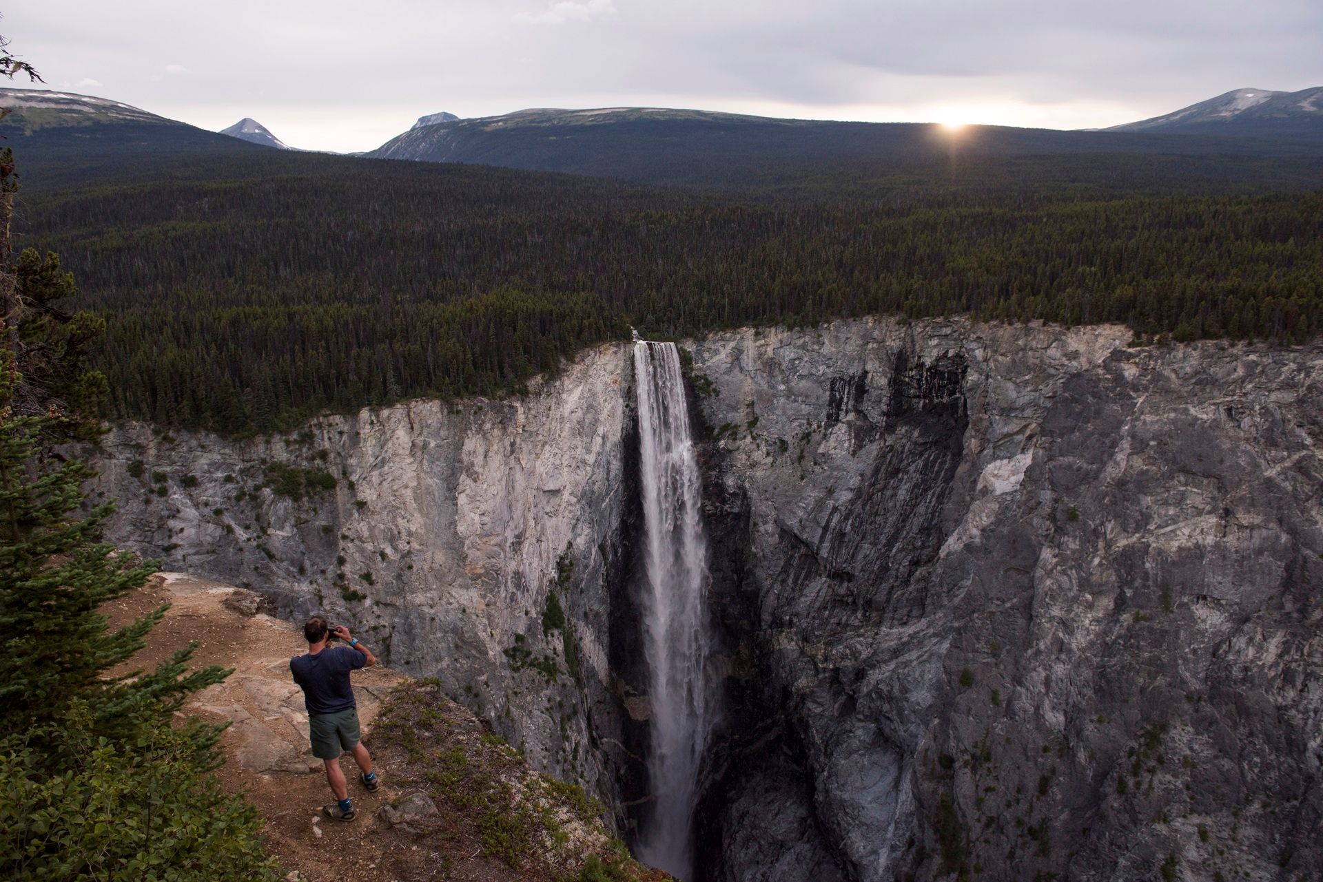 Een persoon staat bij de Hunlen Falls in een rotsachtige kloof, met bos en bergen in de verte.