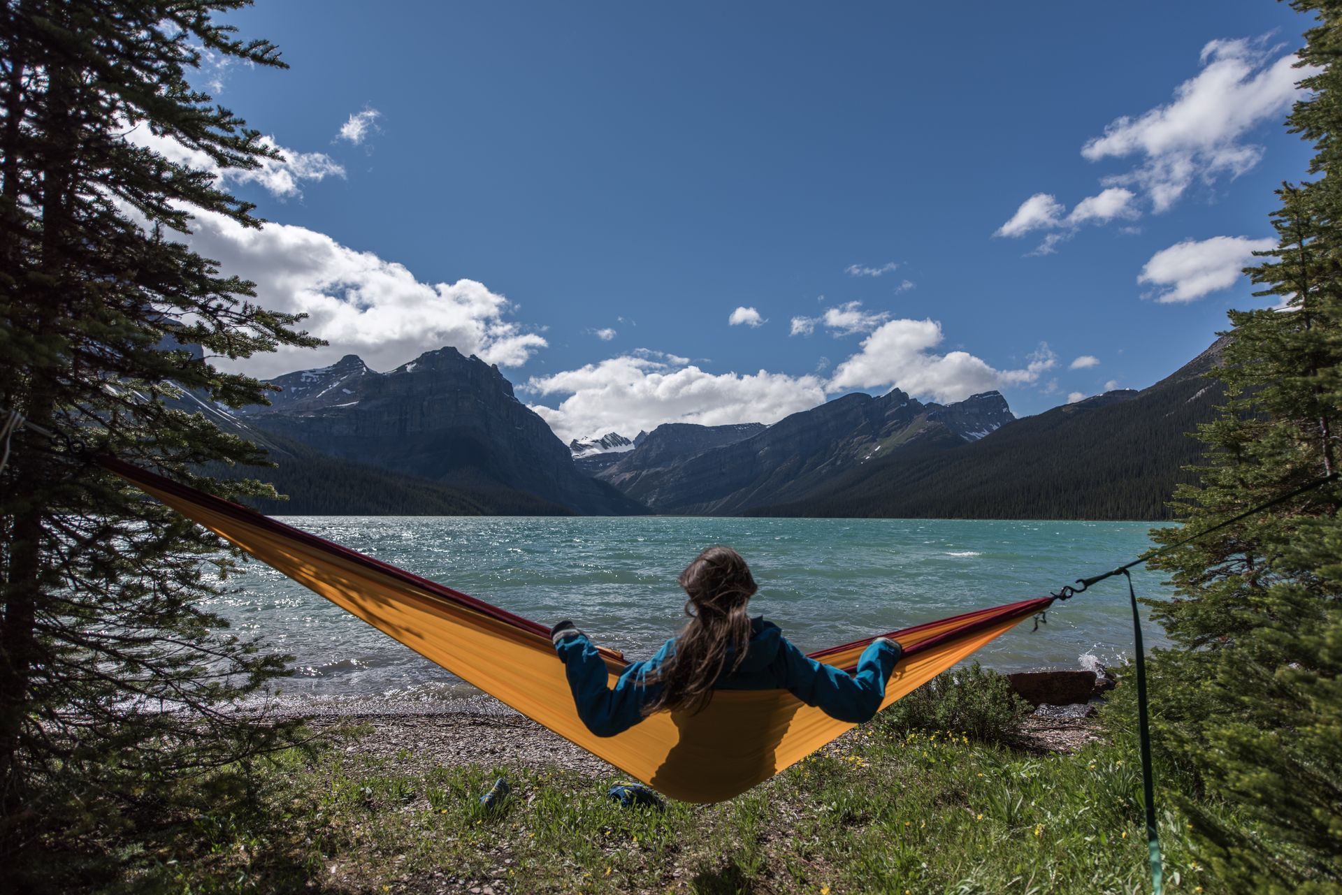 Een persoon ontspant in een oranje hangmat aan Hector Lake aan de Icefields Parkway met bergen en een blauwe lucht op de achtergrond.