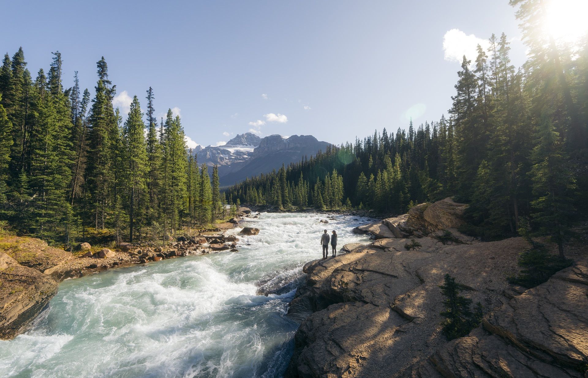 Mistaya Canyon aan de Icefields Parkway met twee mensen op een rotsachtige oever, een dennenbos en besneeuwde bergtoppen in de felle zon.