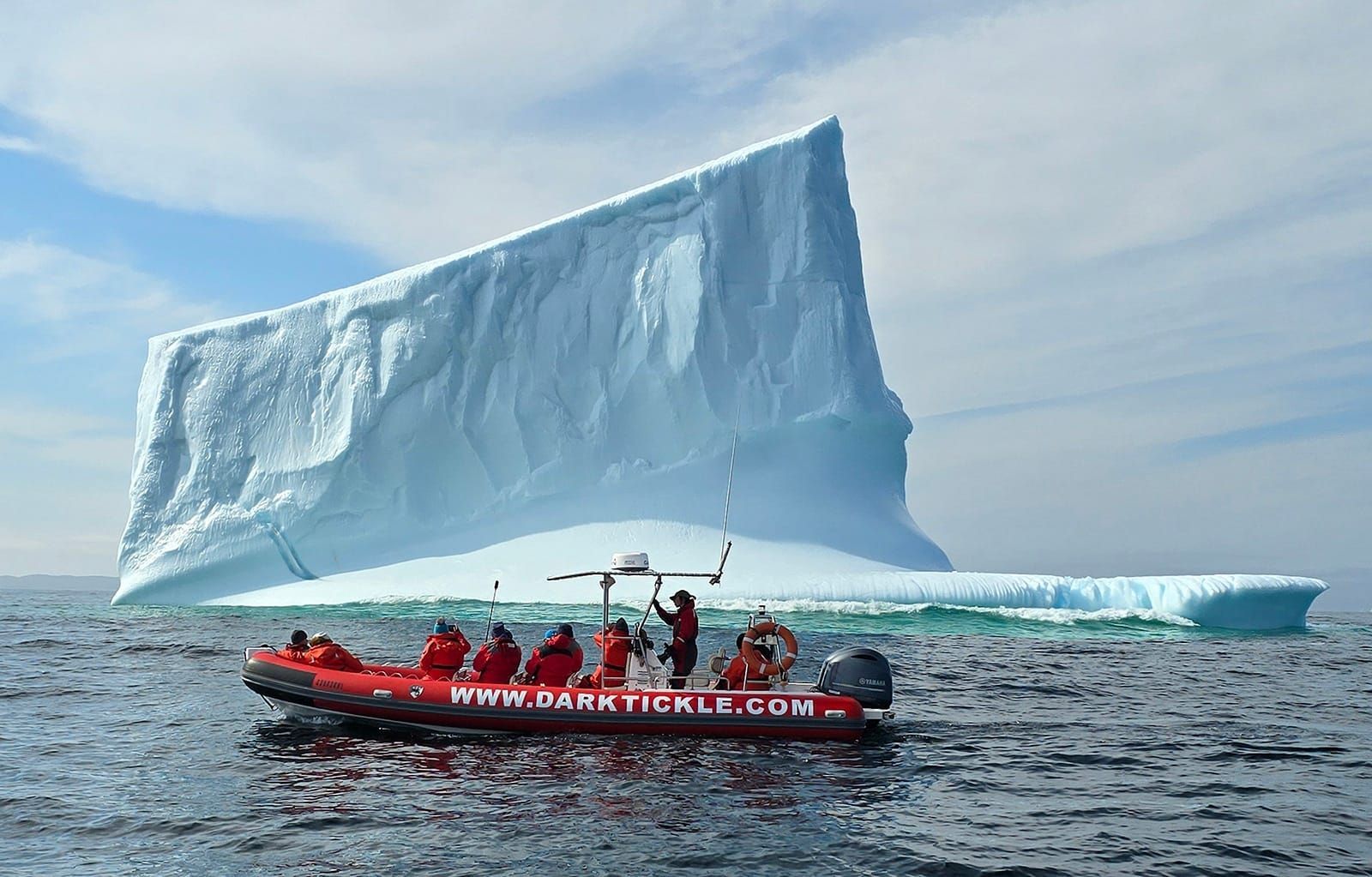 Rode Zodiac boot met mensen aan boord nabij een enorme ijsberg op een kalme oceaan onder een bewolkte hemel in de Atlantische Oceaan van Newfoundland.