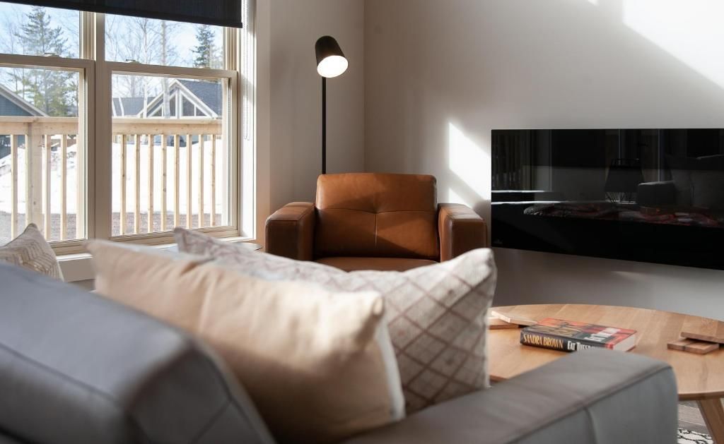 Lichte woonkamer met beige bank, bruine fauteuil, staande lamp, zonlicht door een raam en tv.