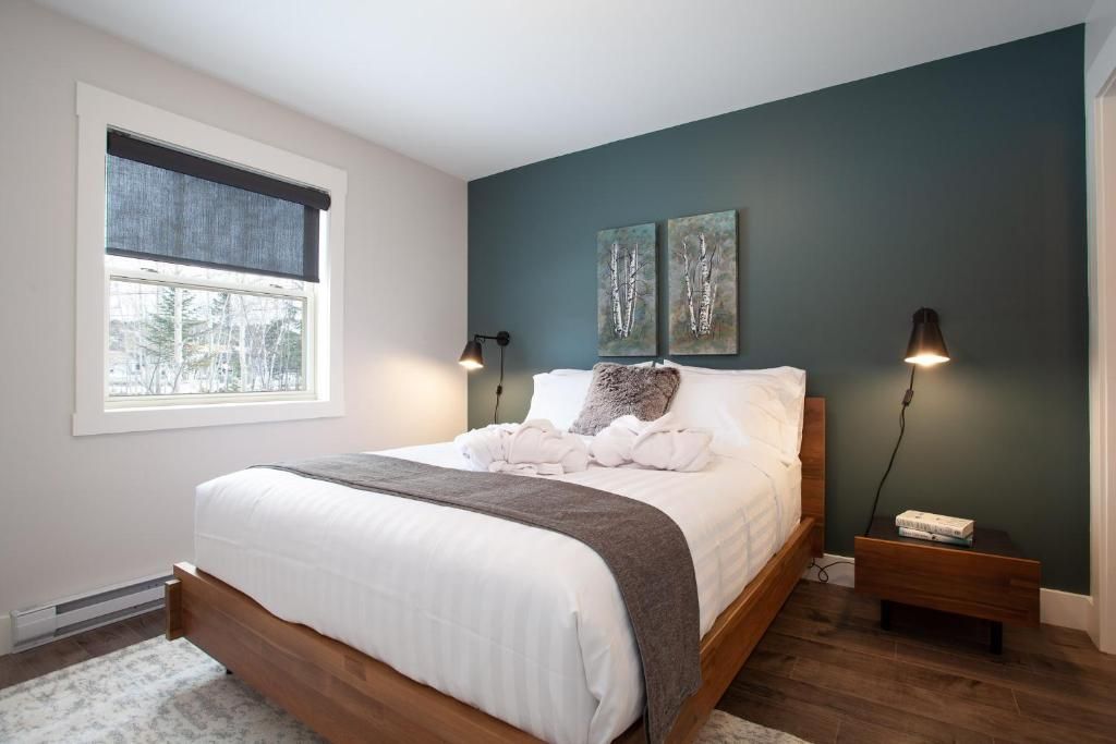 Moderne slaapkamer met wit bed, turquoise accentmuur, houten nachtkastjes en raam met jaloezieën.