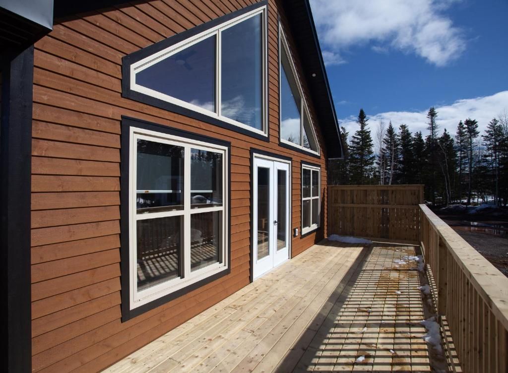 Modern terras bij een huis met houten gevelbekleding, grote ramen en een omheind uitzicht op besneeuwde bomen onder een blauwe hemel.