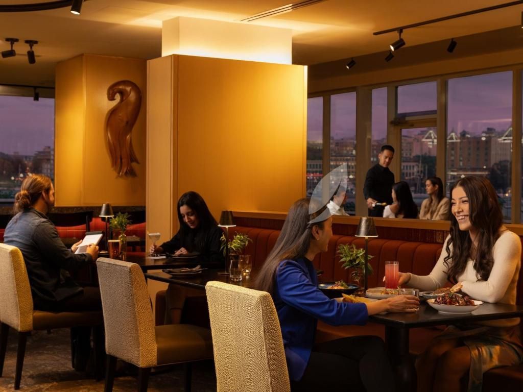 Mensen dineren en praten in een sfeervol verlicht restaurant met grote ramen en uitzicht op de stad.