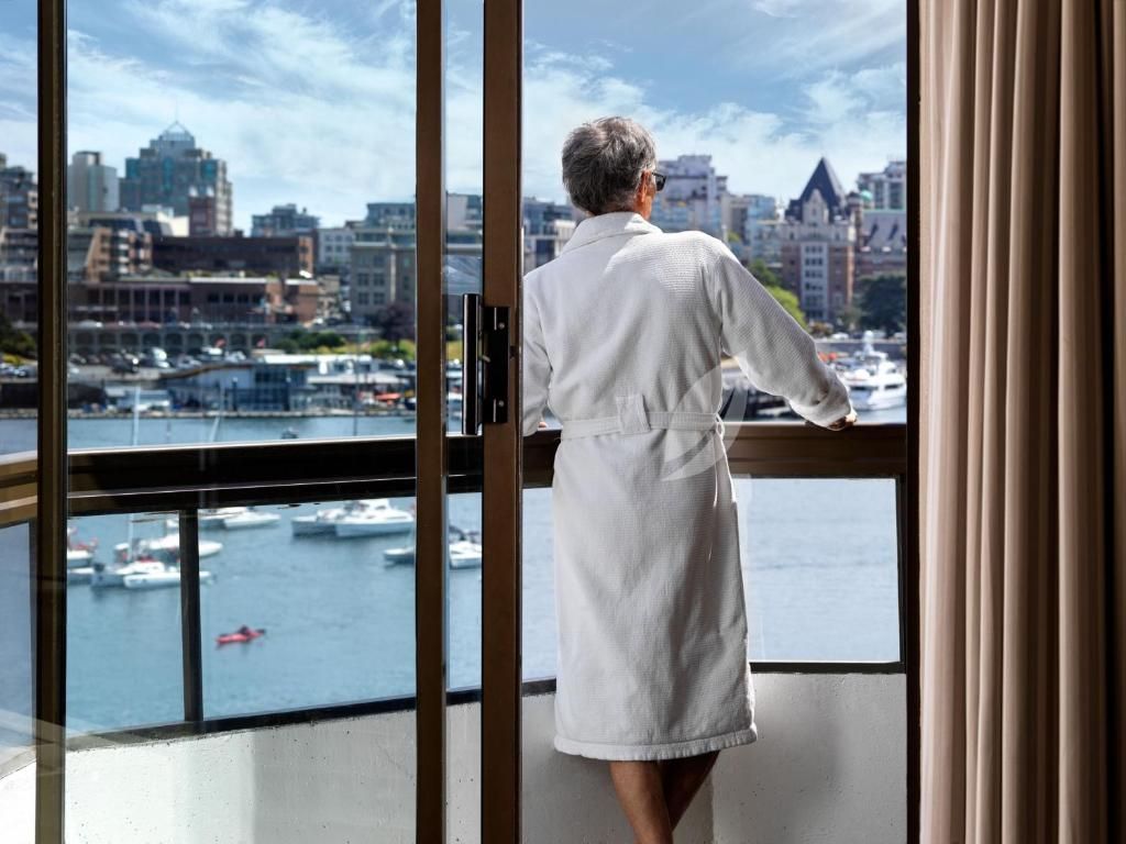 Een persoon in een witte mantel staat op een balkon en kijkt uit over de skyline van een stad aan het water.
