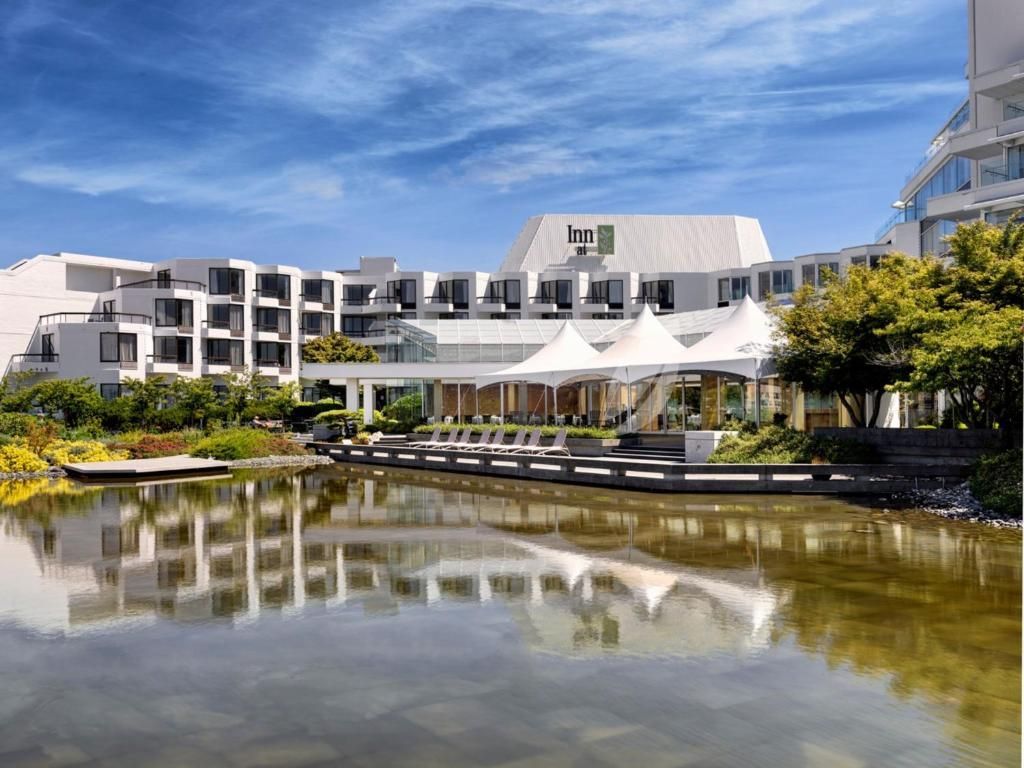 Modern wit hotel aan een vijver, met witte luifels en een helderblauwe lucht die weerspiegeld wordt in het kalme water.