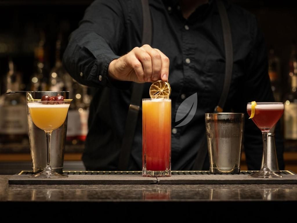 Een barman schenkt achter de bar een gelaagde oranje-rode cocktail in, met nog drie andere drankjes op een rij.