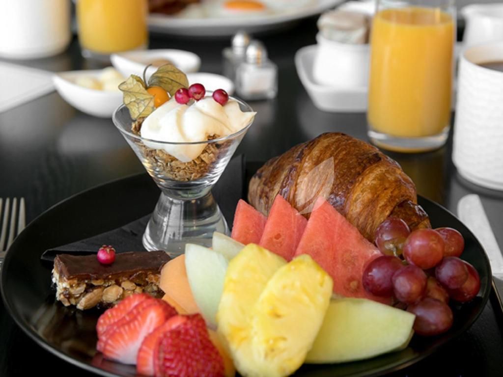 Ontbijtplateau met croissant, fruit, dessertglas en sinaasappelsap op een zwart bord.