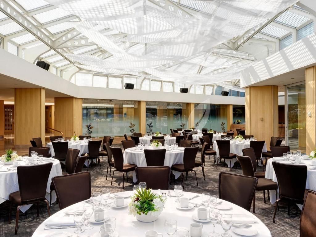 Elegante feestzaal met ronde tafels, donkere stoelen, witte tafelkleden en een helder glazen plafond.