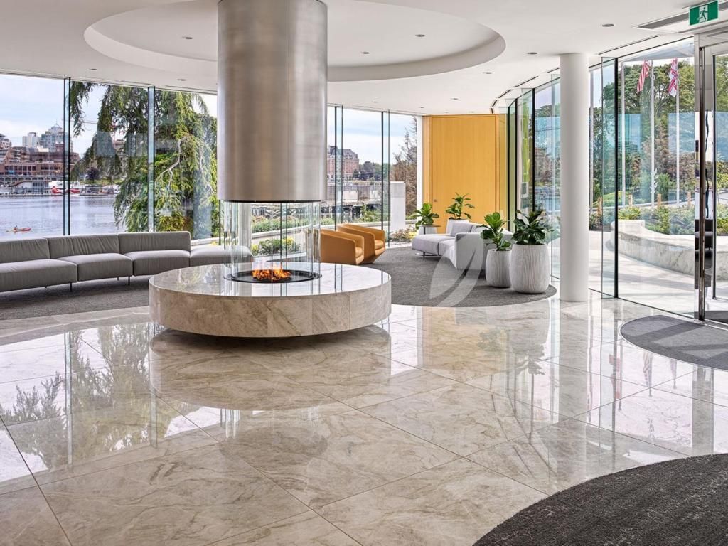 Moderne luxe woonkamer met marmeren vloer, ronde bank, open haard en kamerhoge glazen ramen met uitzicht op het water.