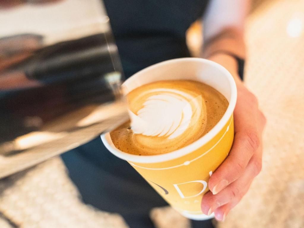 De barista schenkt melk in een geel papieren bekertje voor latte art.