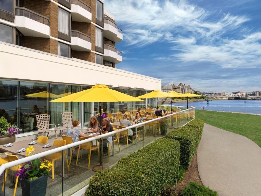 Terras van een restaurant aan zee met gele parasols, naast een modern gebouw en een wandelpad langs het water.