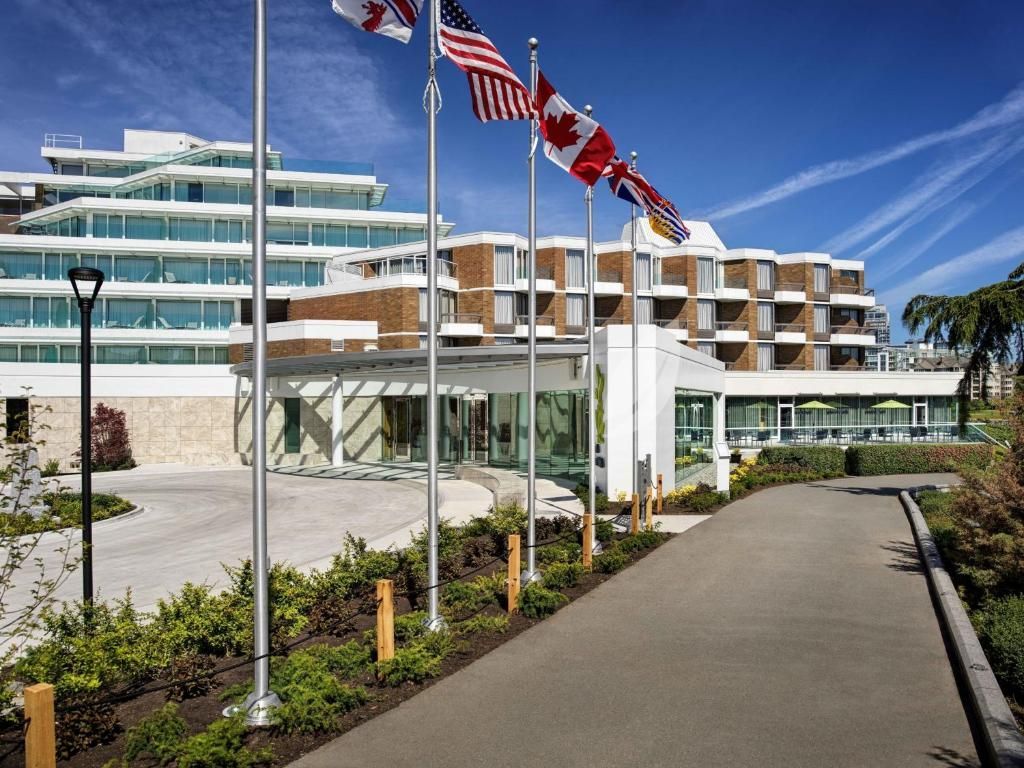 Moderne hotelingang met vlaggenmasten, een gebogen oprit en balkons van blauw glas onder een heldere hemel.