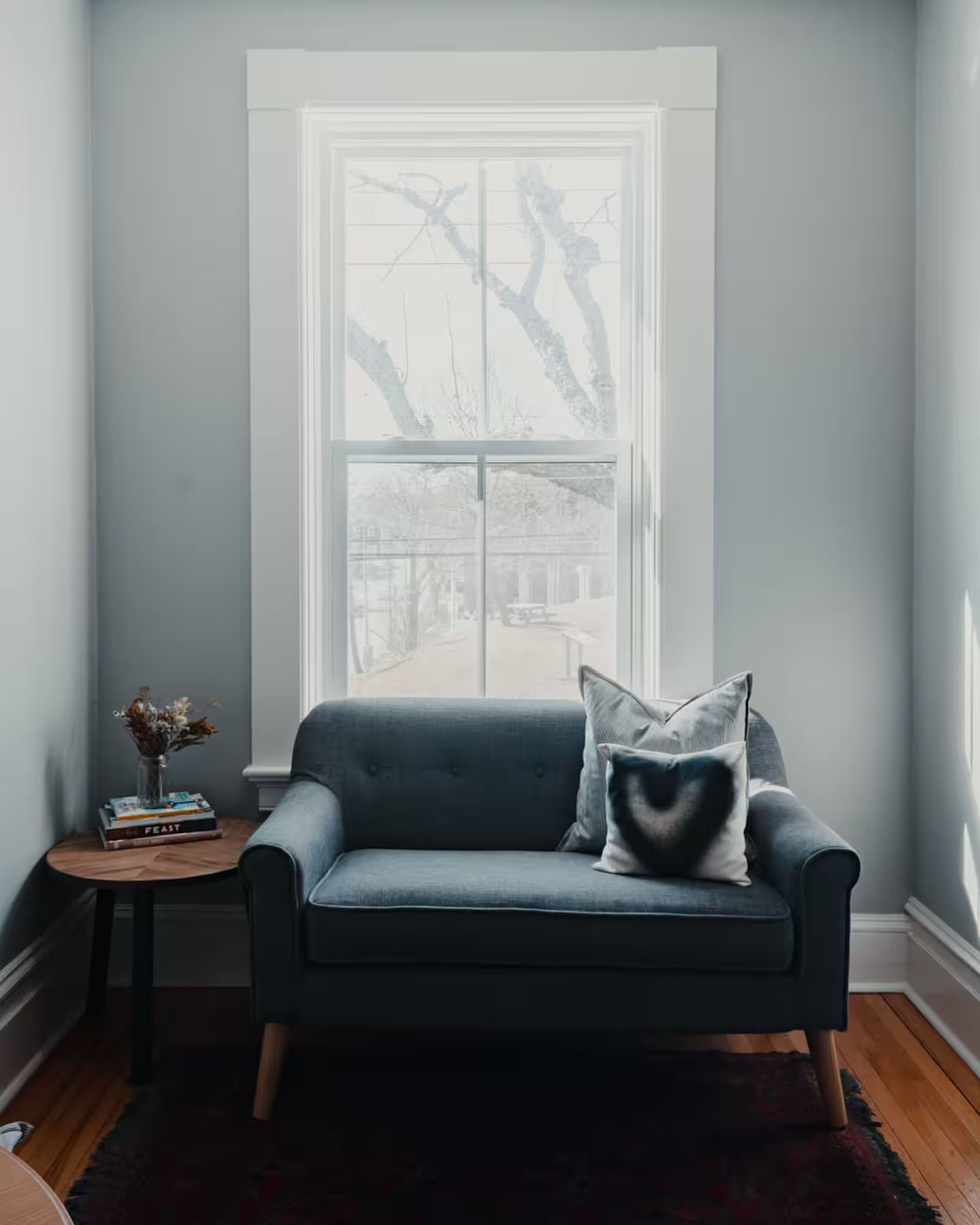 Een grijze fauteuil met een kussen staat bij een licht raam in een minimalistische kamer met een klein bijzettafeltje.
