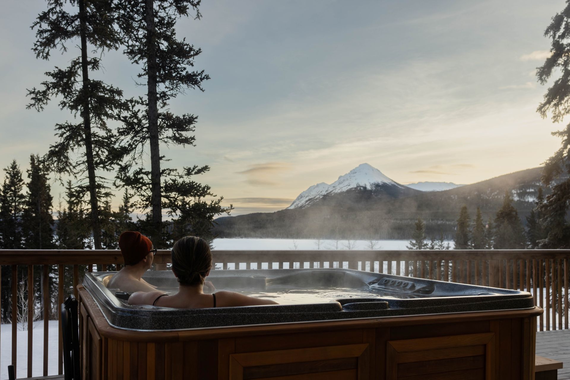 @ExploreCanada Twee mensen ontspannen in een bubbelbad in Yukon met uitzicht op een meer en een besneeuwde berg bij zonsondergang