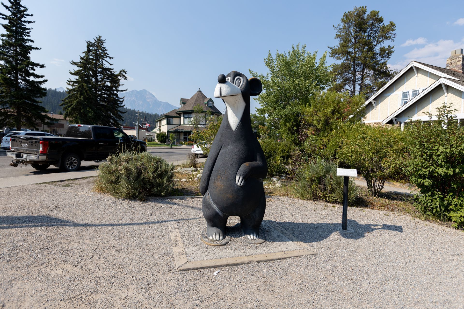 Standbeeld van een zwarte beer in Jasper, Canada langs een grindweg, met bergen, bomen en huizen op de achtergrond.