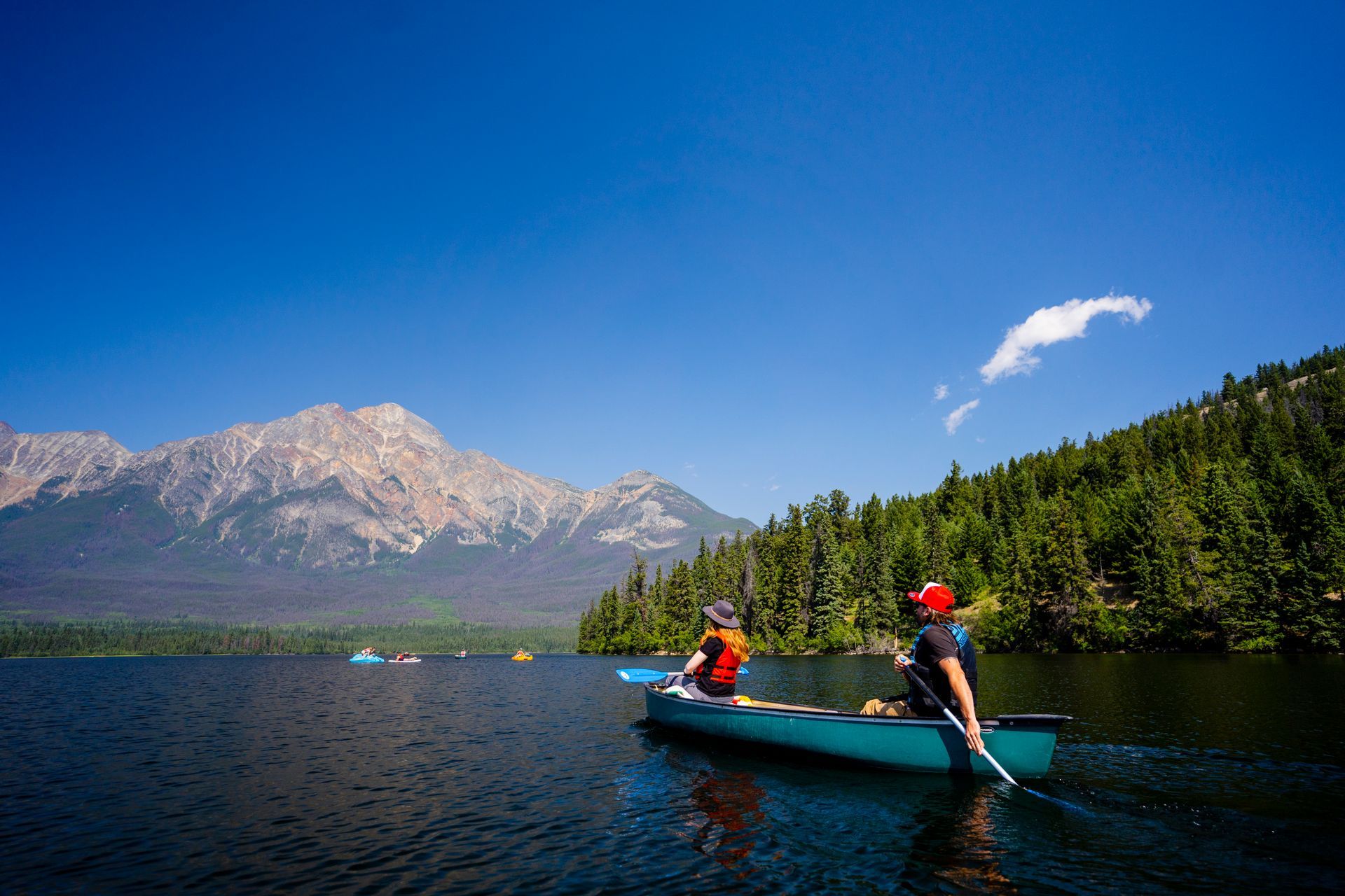 Twee mensen peddelen in een kano op Pyramid Lake in Jasper National Park met dennenbomen en besneeuwde bergtoppen onder een blauwe hemel.