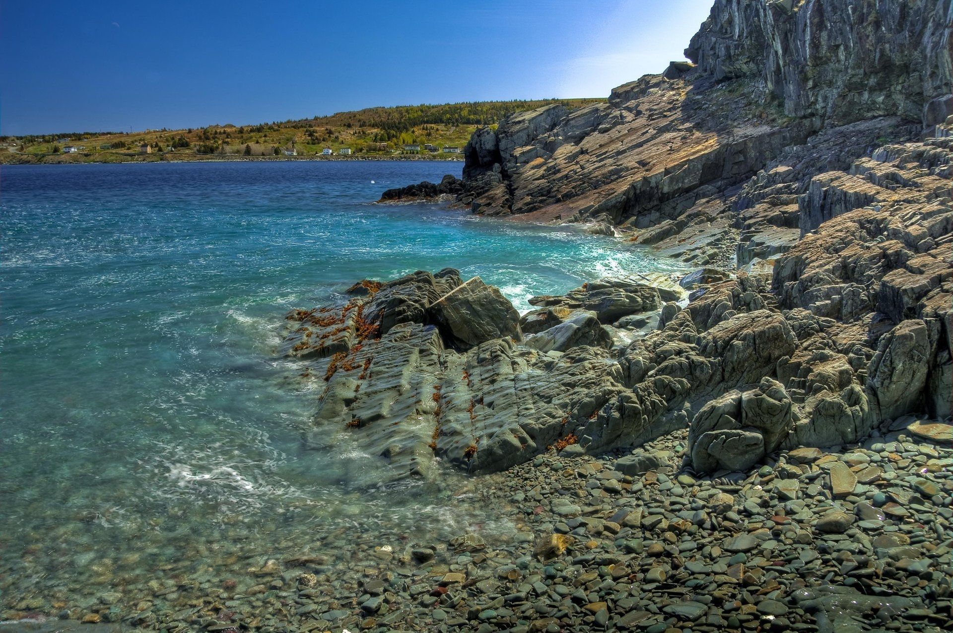 Een rotsachtige kustlijn bij Cupids Newfoundland naast helderblauw water onder een stralende hemel.