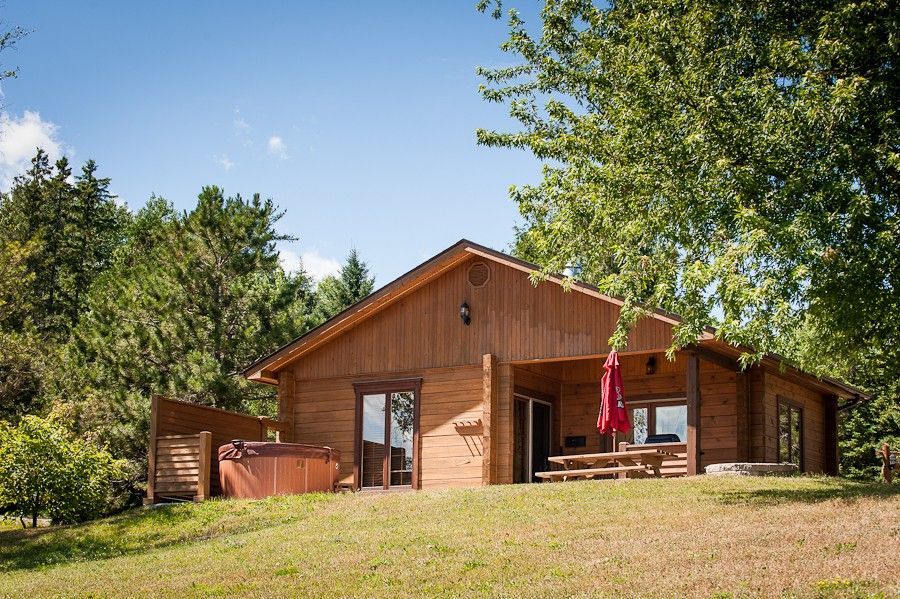 Houten hut met veranda en rode parasol op een zonnige, met gras begroeide heuvel, omgeven door bomen.