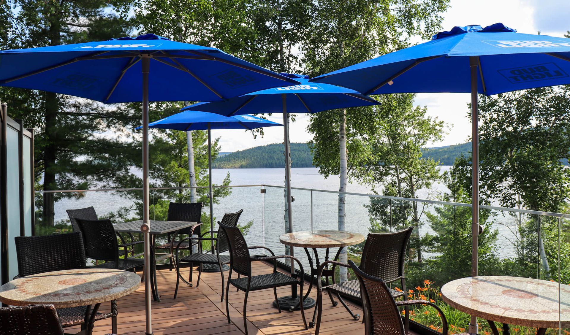 Terras aan het meer met blauwe parasols, tafels en stoelen, met uitzicht op het water en de bomen.