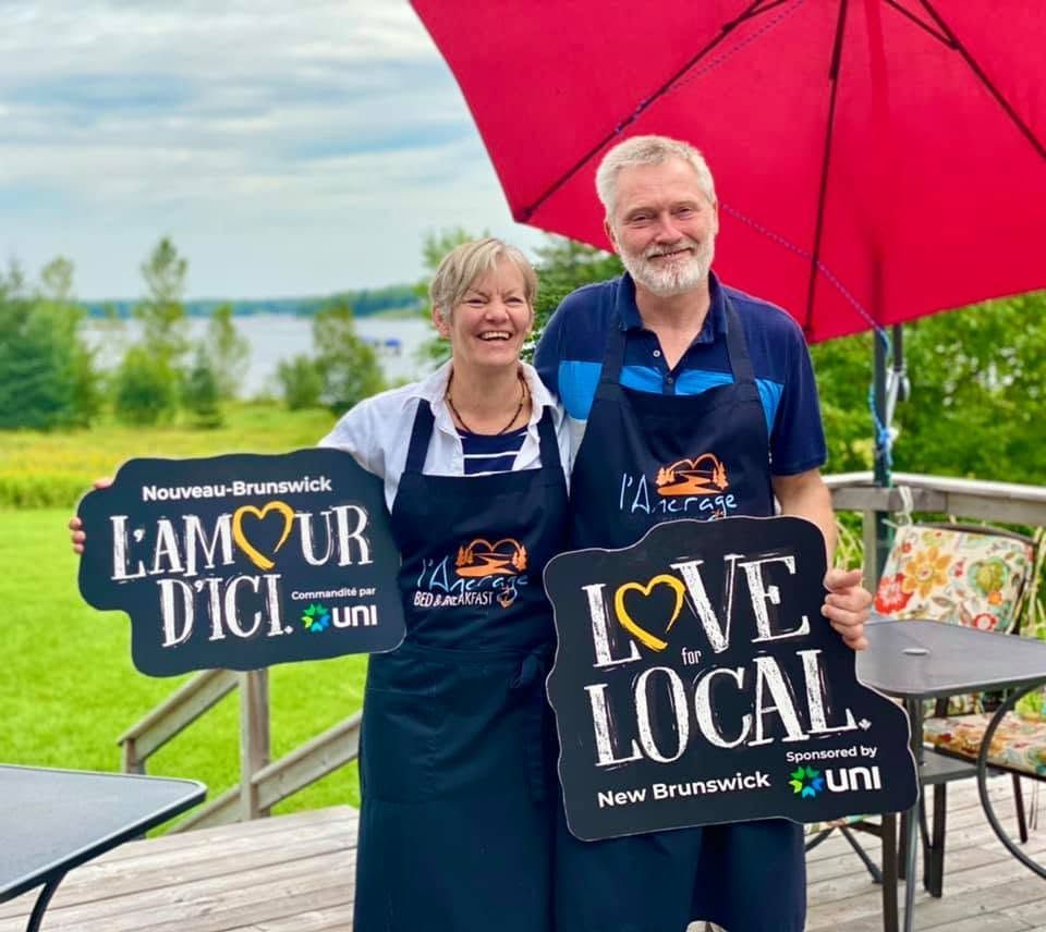 Twee lachende mensen met borden van Team Our Die en Love Local onder een felroze paraplu buiten.