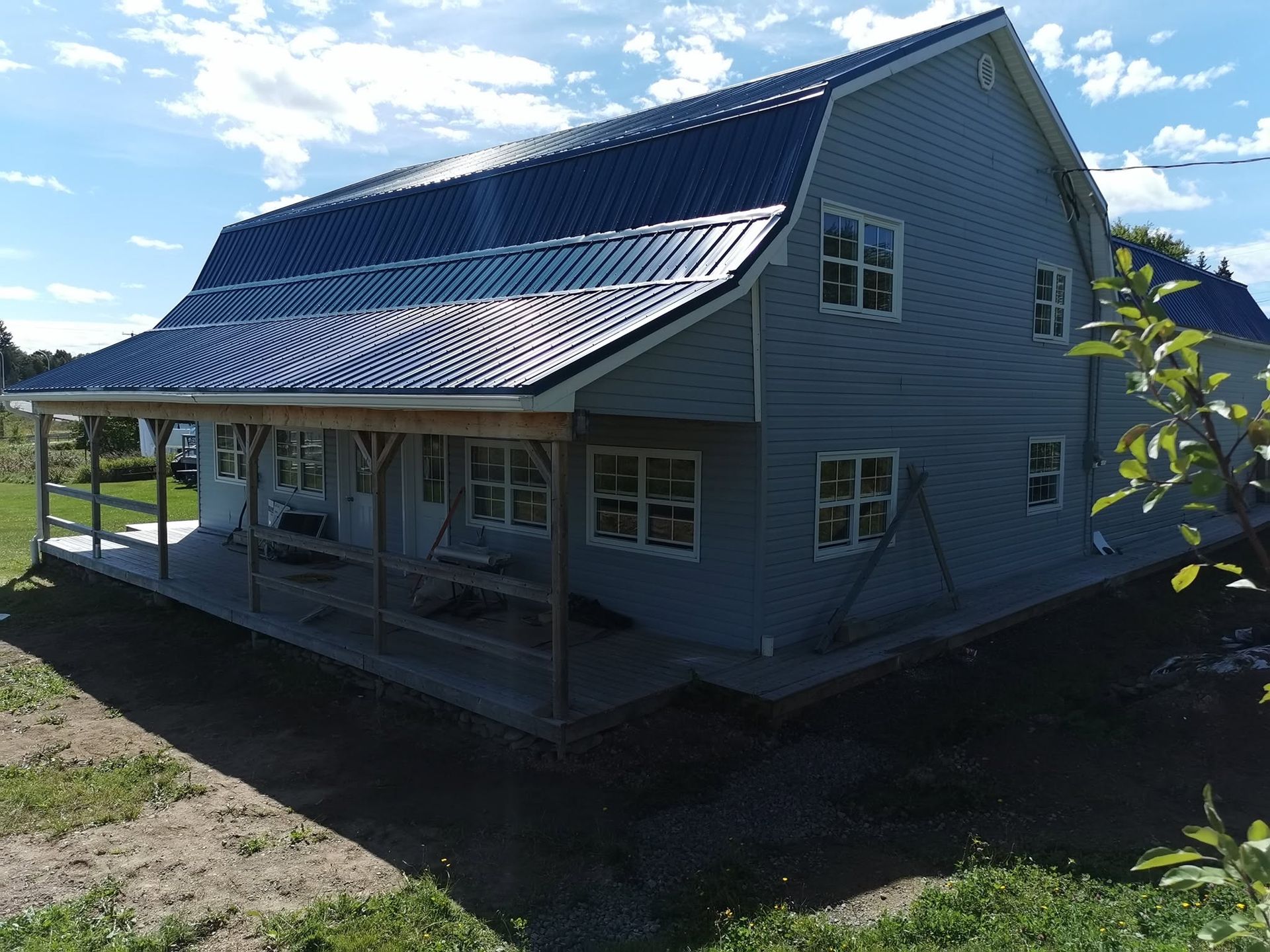 Twee verdiepingen tellend grijs huis met een metalen dak en een overdekte veranda in een zonnige, landelijke omgeving.