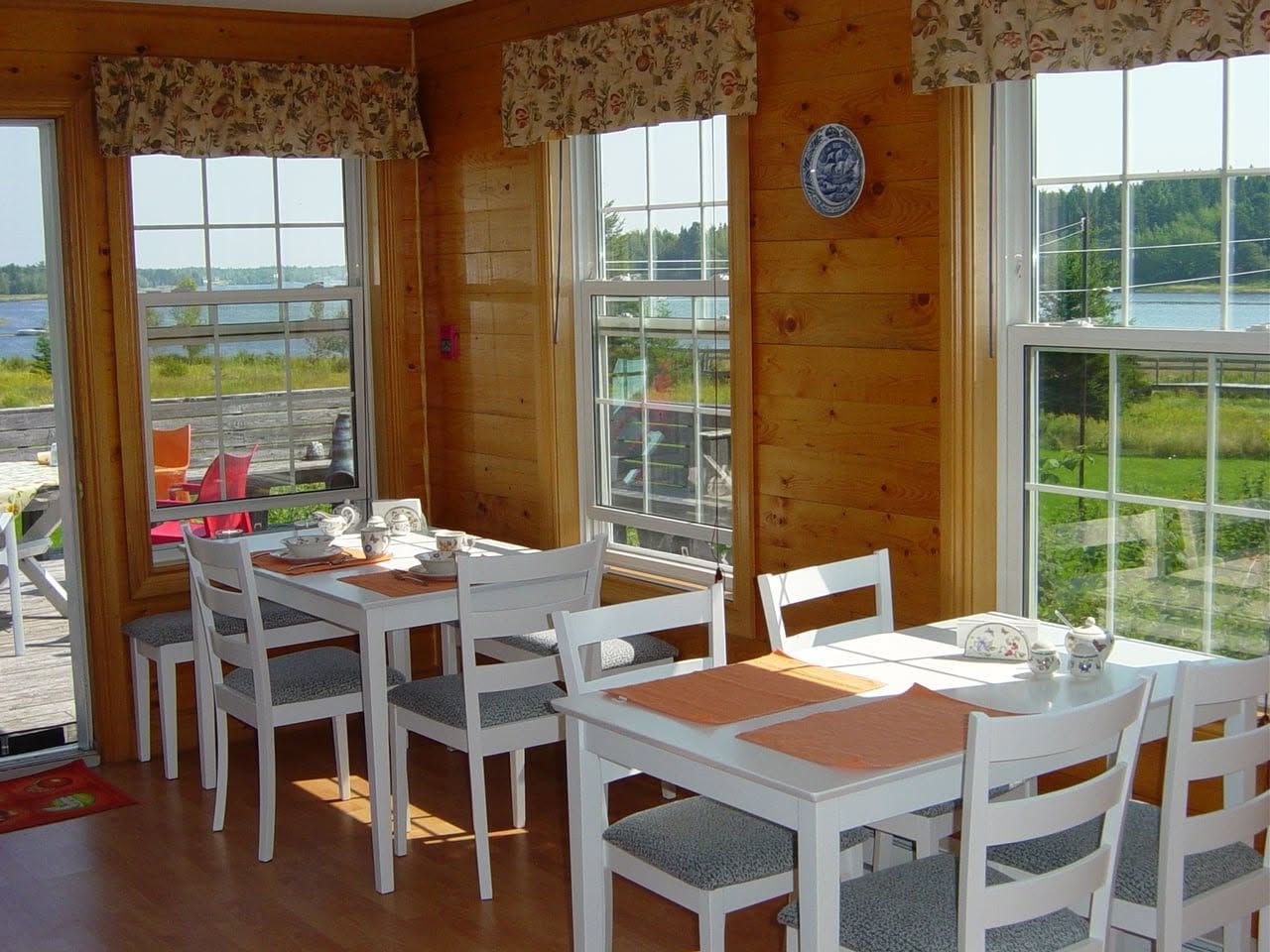 Gezellige, zonnige eetkamer met witte tafels en stoelen bij grote ramen met uitzicht op groene velden.